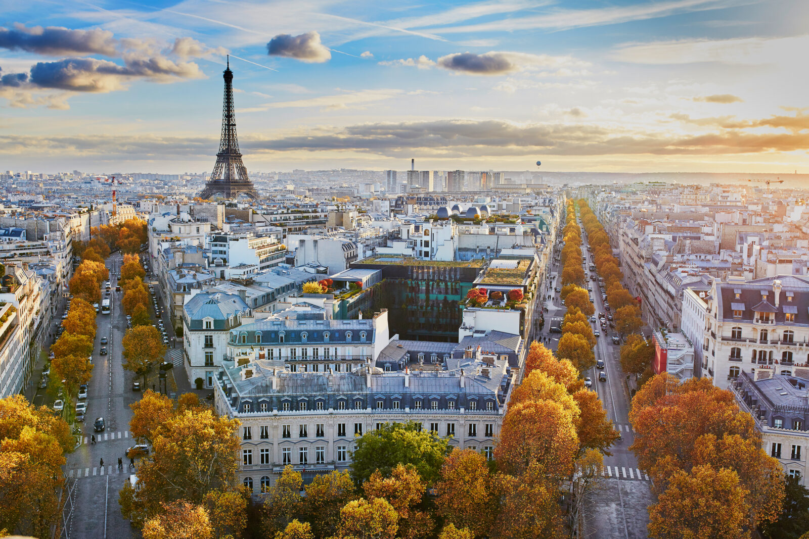 Aerial view of Paris at sunset with the Eiffel Tower in the background, tree-lined streets, and historic buildings with autumn-coloured leaves.