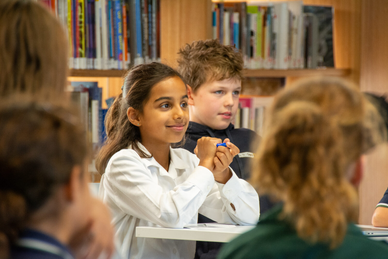 A girl in a white shirt sits at a table in a library, smiling and holding a pen, surrounded by other children. Bookshelves filled with books are visible in the background.