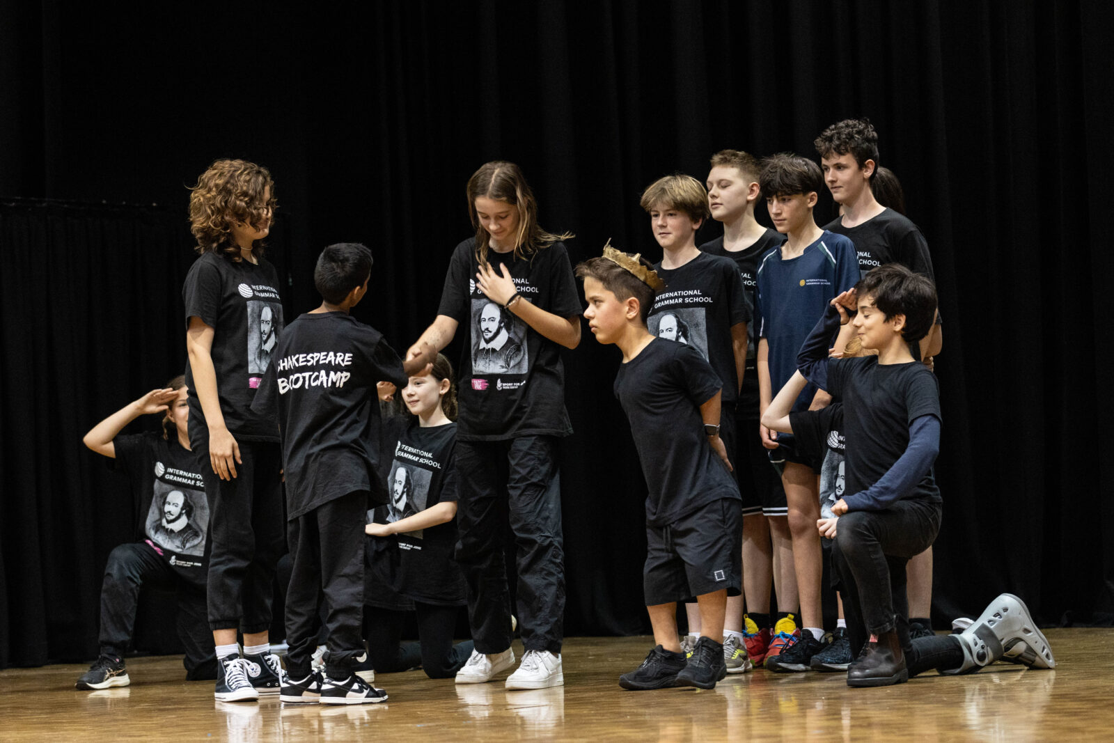 A group of children and teenagers in black Shakespeare Bootcamp T-shirts perform on stage, some standing and some kneeling, with dramatic expressions and gestures against a dark background.