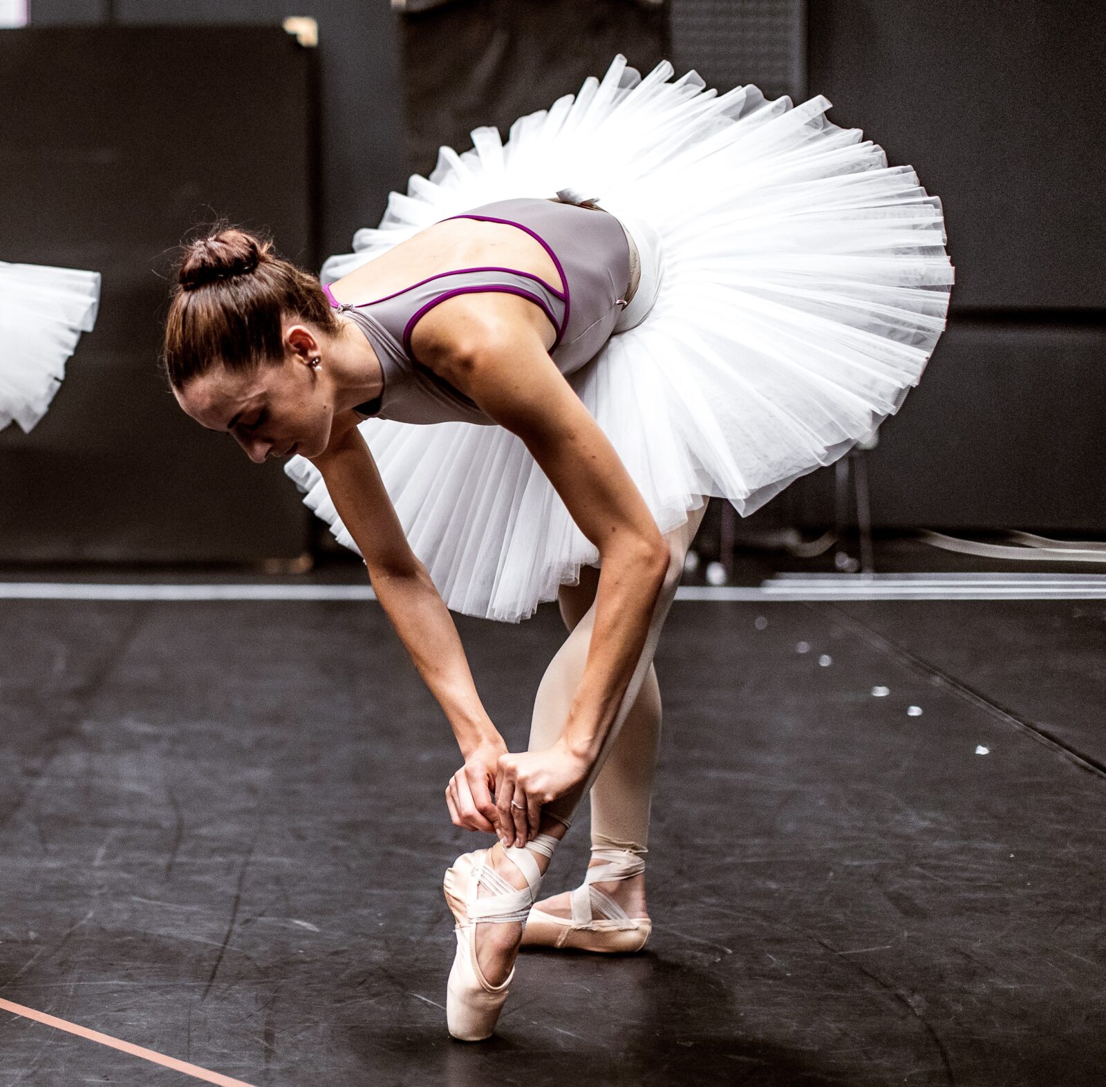 A ballerina in a white tutu bends forward to adjust her pointe shoe on a dark dance studio floor, her hair in a bun and her arms reaching down gracefully.