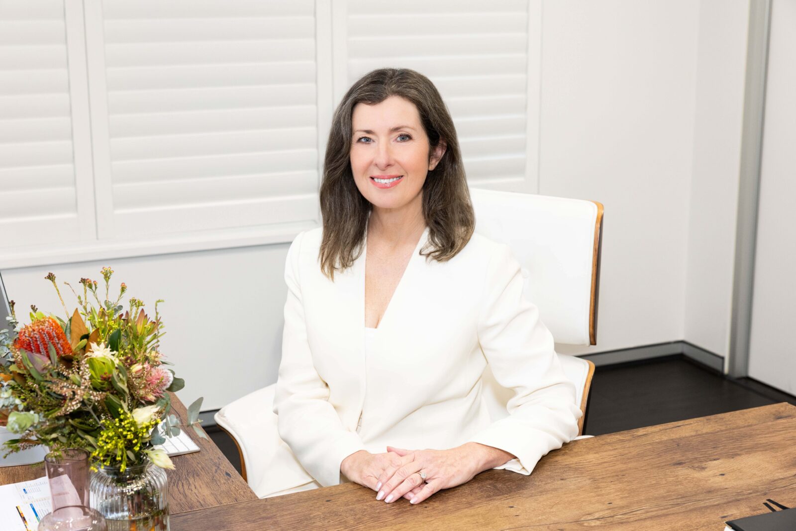 Shauna Colnan, IGS Principal, sits at a wooden desk in a white office, wearing a white blazer and smiling. A bouquet of flowers and papers are on the desk in front of her.