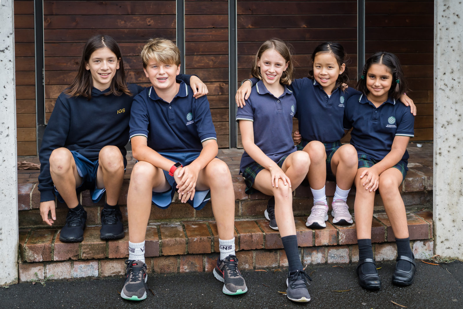 Five school children sit together on brick steps, smiling at the camera. They wear navy blue IGS uniforms and trainers, and have their arms around each other. The background features wooden panelling.