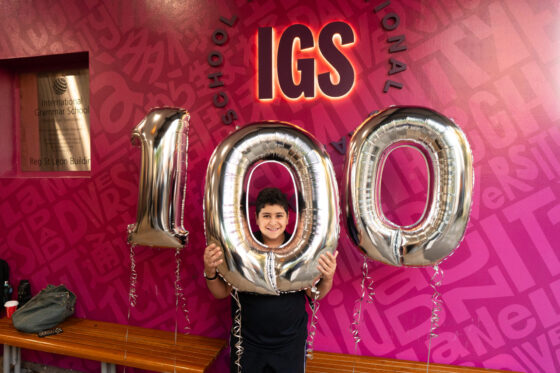 A smiling person holds large silver balloons forming 100 in front of a pink wall with IGS and school-related text.