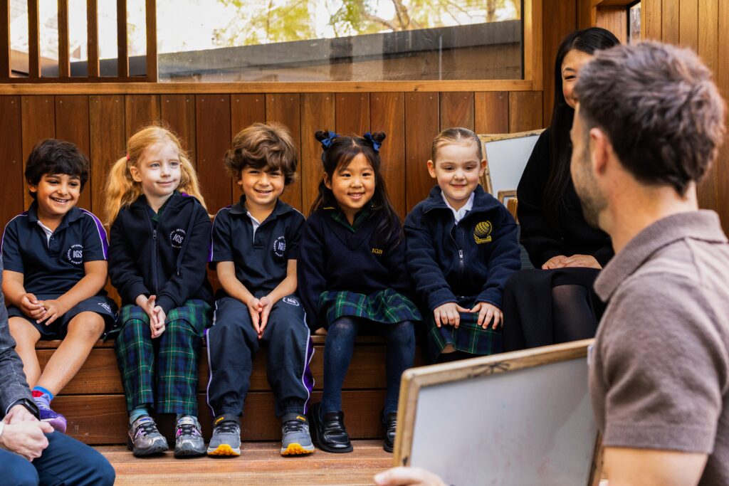 Five young children in school uniforms sit on wooden steps, smiling and listening to an adult holding a whiteboard. The setting appears to be a classroom or outdoor learning area.