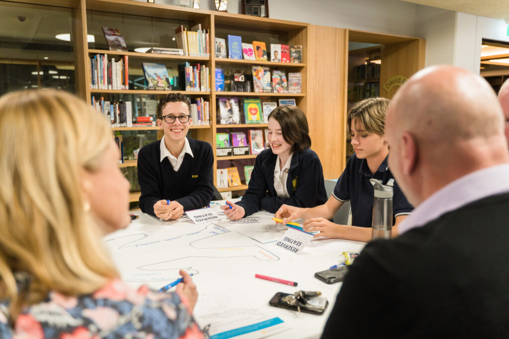 A group of students and adults sit around a table in a library, collaborating and smiling whilst working on a large sheet of paper with felt-tip pens. Bookshelves filled with books are visible in the background.