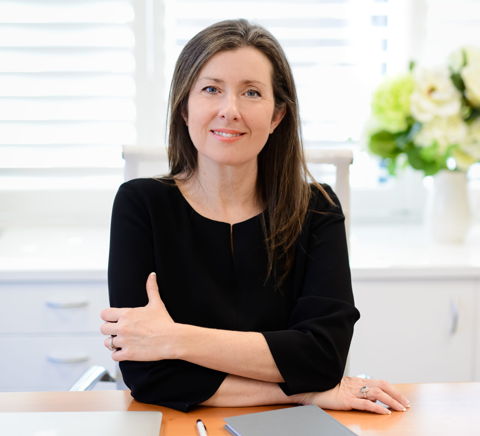 Shauna Colnan, IGS Principal, with long brown hair and a warm smile, sits at a desk with her arms folded. A notebook, pen, and laptop sit before her; white blinds and flowers brighten the background.