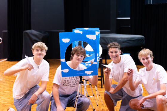 Four smiling teenagers in white shirts kneel indoors, each holding a paper aeroplane. They pose with a blue board decorated with clouds and a circular cut-out, suggesting an engaging enterprise activity or event for future business leaders.