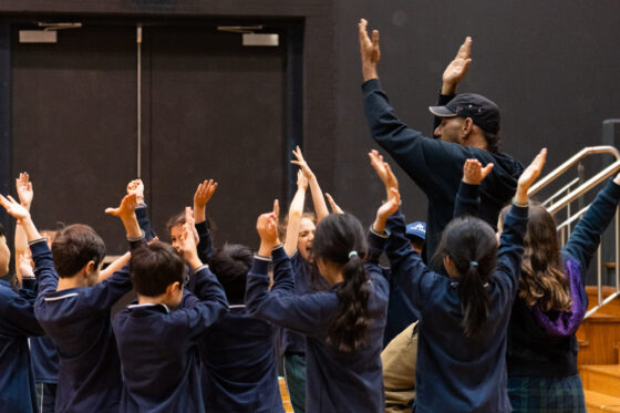 A group of children and an adult instructor raise their arms together in a gymnasium, facing away from the camera, engaging in an energetic group activity.