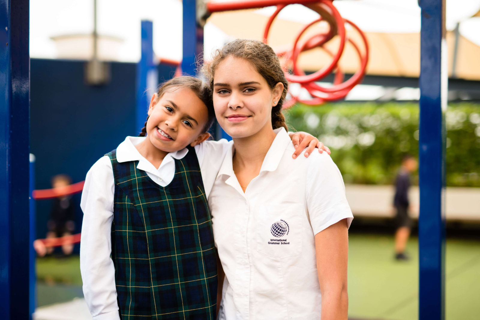 Two girls in school uniforms stand arm-in-arm, smiling at the camera on a playground. The younger wears a checked dress and white shirt; the older, in a logo shirt, shares how bursaries make their school experience possible. Playground equipment is in the background.